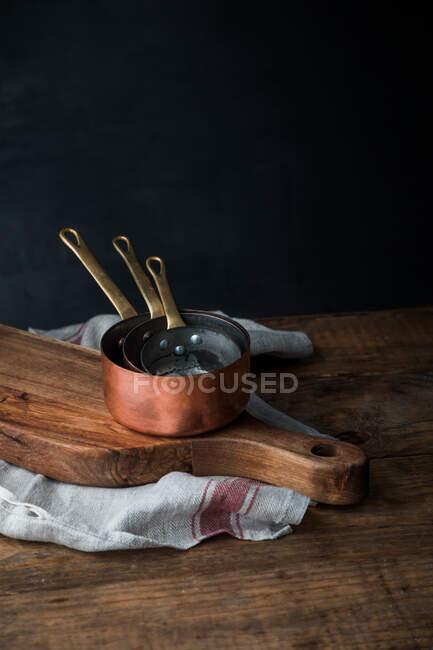 Stack of copper saucepans placed on wooden cutting board and linen towel on rustic table on gray background — Stock Photo