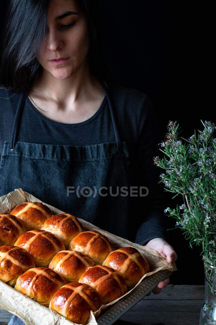 Panadero femenino sosteniendo hojaldre recién horneado panecillos cruzados calientes en bandeja para hornear - foto de stock