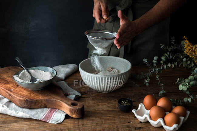Unrecognizable uomo setacciando farina in ciotola mentre si prepara pasticceria in cucina rustica a casa — Foto stock