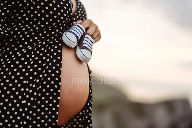Cropped unrecognizable pregnant female holding little shoes on belly while standing against blurred countryside landscape — Stock Photo