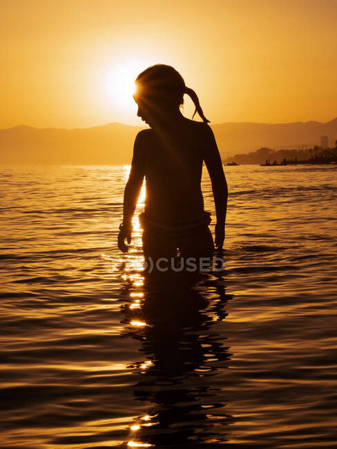 Silhouette di bambina anonima in piedi in acqua di mare contro la luce del tramonto in estate — Foto stock