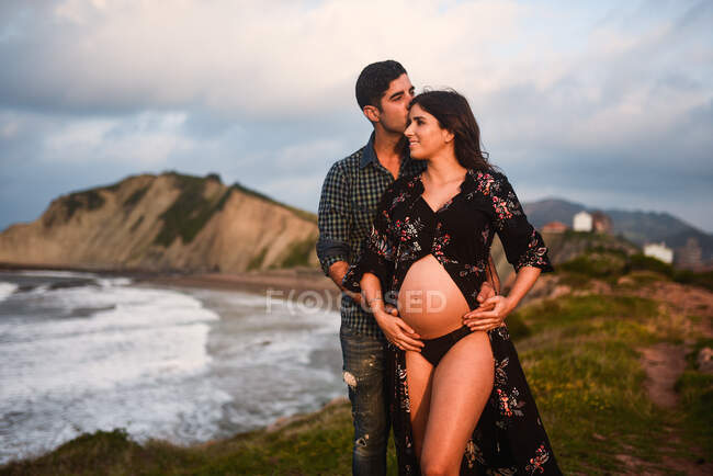 Loving man embracing and kissing happy pregnant wife dressed in stylish outfit revealing tummy while standing together on seashore against mountains — Stock Photo