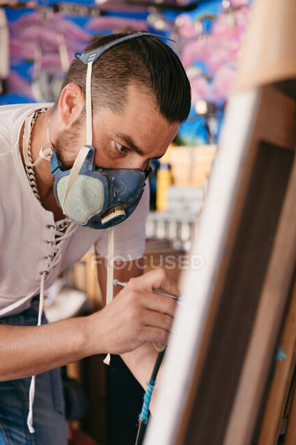 Männlicher Künstler in Atemschutzmaske mit Spritzpistole, um während der Arbeit in der Kreativwerkstatt Bild auf Leinwand zu malen — Stockfoto