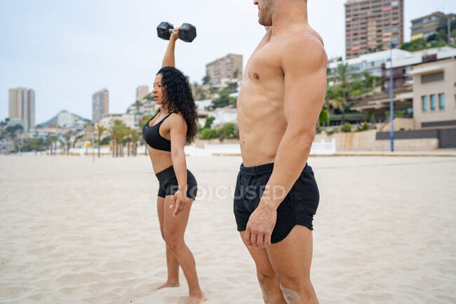 Вид сбоку мускулистых мультиэтнических спортсменов, тренирующихся с гантелями во время тренировок на песчаном пляже летом — стоковое фото