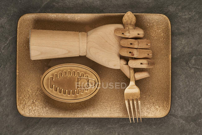 Искусственная деревянная рука с вилкой, расположенная на золотом подносе возле герметичной банки с консервами на столе — стоковое фото
