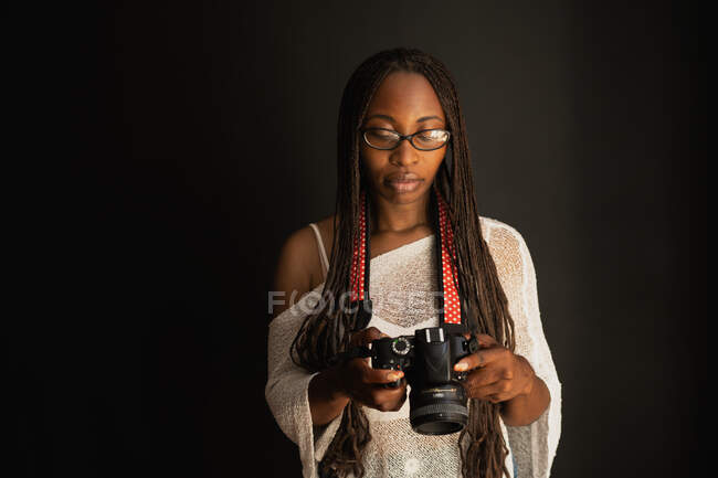 Grave fotógrafa afroamericana con trenzas mirando a través de fotos tomadas en cámara profesional mientras está de pie sobre fondo negro - foto de stock