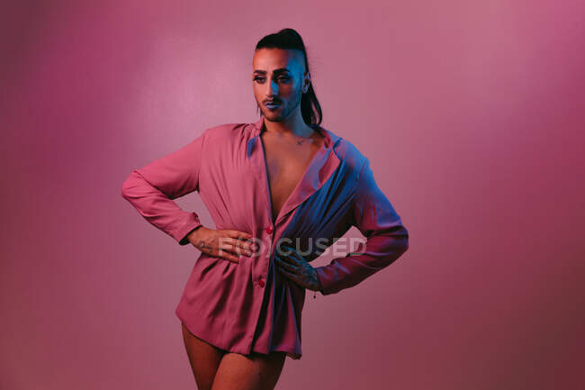Retrato de mujer barbuda transgénero glamorosa en maquillaje sofisticado posando con las manos en la cintura sobre fondo rosa en el estudio mirando hacia otro lado - foto de stock