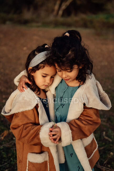 Милые мирные сестрички в одинаковой верхней одежде обнимают друг друга, стоя вместе на размытом фоне природы. — стоковое фото