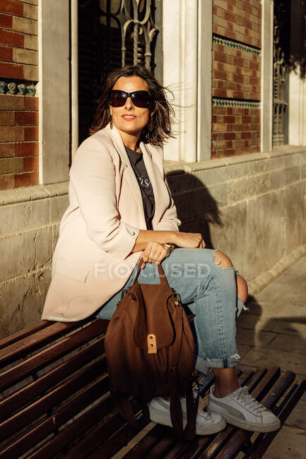 Corps complet de femme à la mode dans une tenue décontractée élégante et des lunettes de soleil avec sac à main assis sur le banc et profiter de la lumière du soleil tout en se reposant sur la rue urbaine — Photo de stock