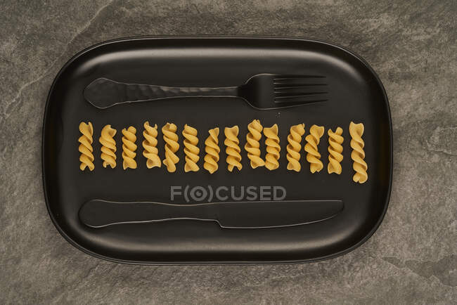 Top view of black fork and knife placed near uncooked fusilli pasta on tray on table — Stock Photo