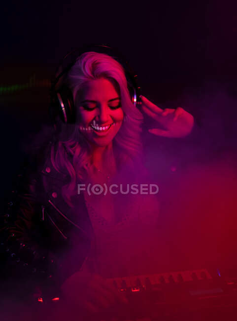 Fröhliche junge DJ-Frau mit Kopfhörer lächelt und benutzt Synthesizer, um Musik zu spielen, während sie während einer Party in einem Nachtclub im Rauch steht — Stockfoto