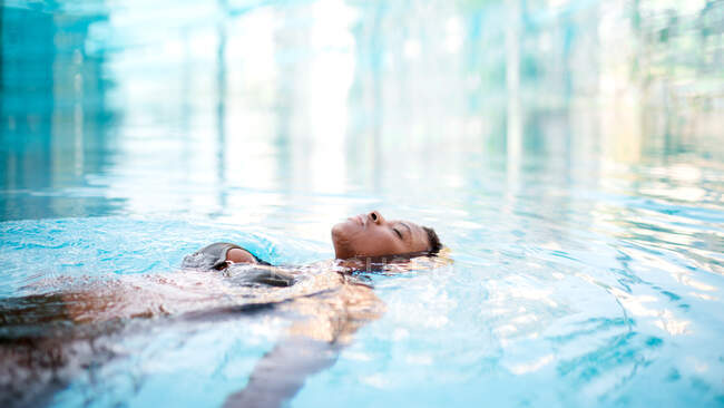 Tranquille femme afro-américaine en bikini couchée sur l'eau claire dans la piscine et profitant de l'été avec les yeux fermés — Photo de stock