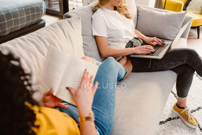 Анонимная лесбийская пара просматривает нетбук и читает интересную книгу, отдыхая на удобном диване в современной гостиной — стоковое фото