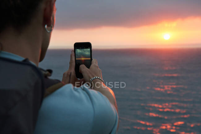 Rückansicht eines unkenntlich gemachten männlichen Reisenden, der im Sommer Sonnenuntergänge über dem Meer mit dem Smartphone fotografiert — Stockfoto