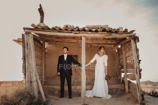 Homem e mulher de corpo inteiro em roupas elegantes de mãos dadas em pé em abrigo de madeira resistido no dia do casamento no Parque Natural Bardenas Reales em Navarra, Espanha — Fotografia de Stock