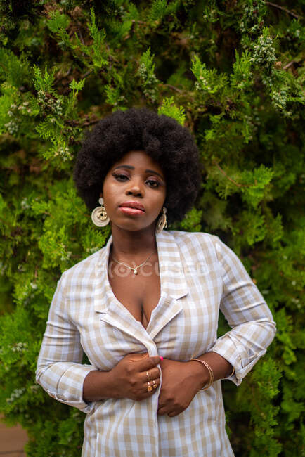 Mulher afro-americana jovem elegante com cabelo afro e brincos elegantes em pé entre folhas verdes no jardim e olhando para a câmera — Fotografia de Stock