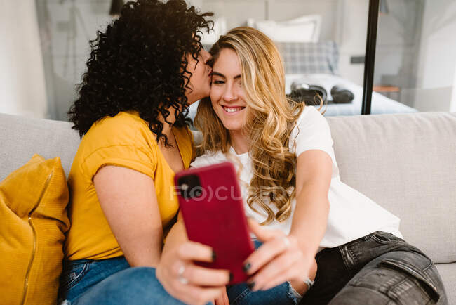 Crop романтична лесбіянка пара в повсякденному одязі цілується один з одним і приймає селфі на смартфон, сидячи на затишному дивані — стокове фото