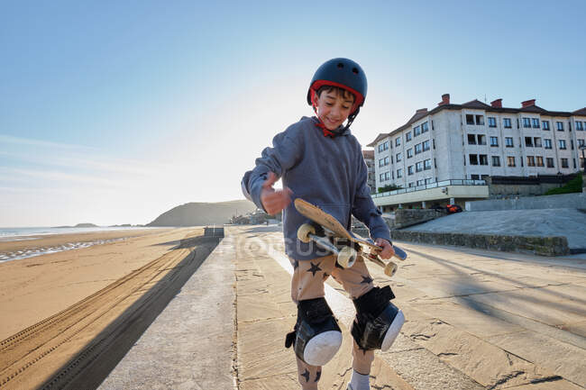 Fröhlicher Teenager mit Helm steht im Sommer mit Skateboard auf Promenade am Meer — Stockfoto