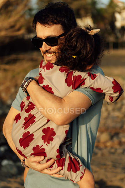 Vue latérale de l'homme adulte en lunettes de soleil portant et embrassant fille avec les yeux fermés le week-end dans la campagne — Photo de stock