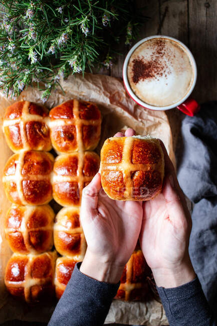 From above crop person hand holding bun on background of table with tasty lush freshly baked hot cross buns and cup of coffee — Stock Photo