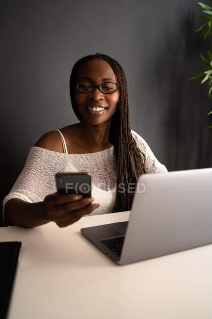 Fröhliche afroamerikanische Freelancerin sitzt am Tisch am modernen Arbeitsplatz und surft auf dem Handy, während sie fernab von zu Hause an einem Projekt arbeitet — Stockfoto