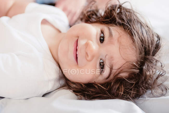 Adorável sorridente menina com olhos castanhos e cabelos cacheados deitado na cama com pai colheita e olhando para a câmera — Fotografia de Stock