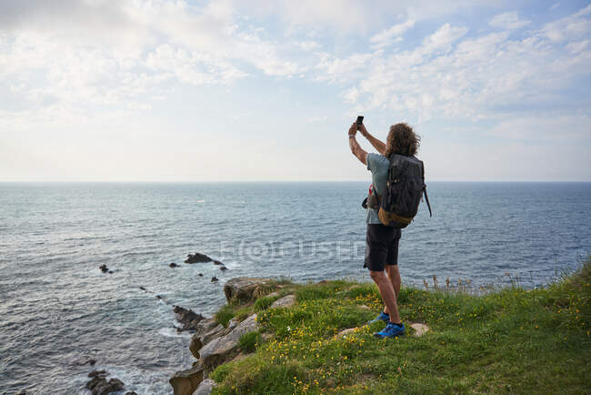 Назад чоловічий піший турист робить самостріл на смартфоні, стоячи на пагорбі на фоні моря під час походу влітку — стокове фото