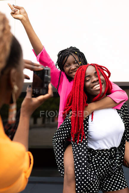 Crop African American female taking picture on photo camera of laughing girlfriend giving piggyback to best friend with hand raised — Stock Photo