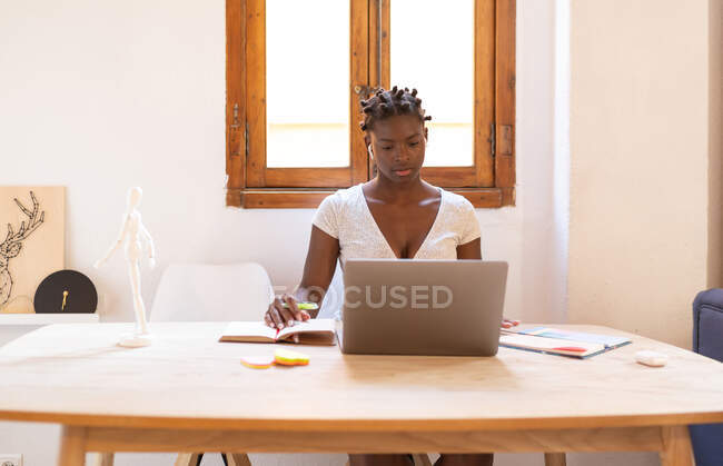 Afroamerikanische Freiberuflerin macht sich Notizen im Notizbuch, während sie an einem Projekt arbeitet und zu Hause mit Laptop am Tisch sitzt — Stockfoto