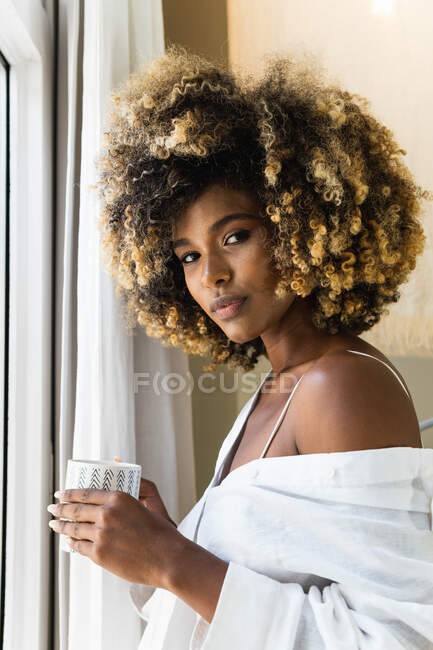 Friedliche Afroamerikanerin mit lockigem Haar trägt ein weißes Hemd, das am Fenster steht und morgens eine Tasse Getränk trinkt — Stockfoto