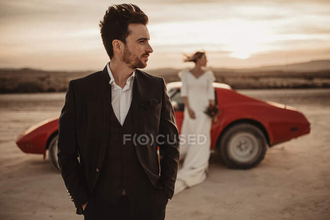 Uomo in giacca e cravatta distogliendo lo sguardo mentre in piedi contro auto sportive rosse e sposa al tramonto nel Parco Naturale Bardenas Reales in Navarra, Spagna — Foto stock