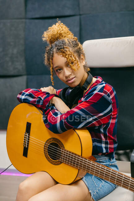 Jovem guitarrista afro-americana com guitarra clássica e fone de ouvido sentado com braços cruzados em poltrona enquanto olha para a câmera — Fotografia de Stock