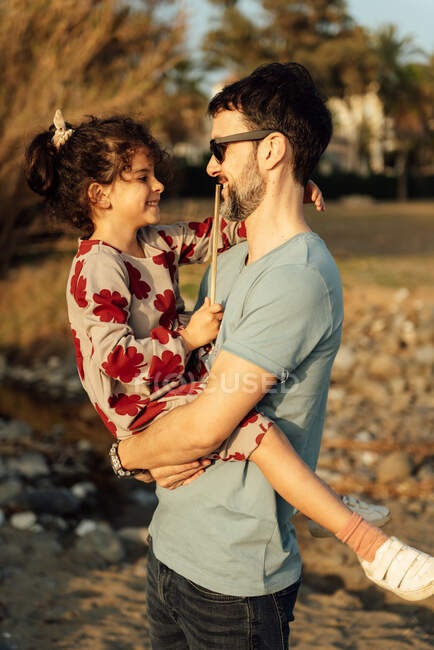 Vista lateral del hombre adulto en gafas de sol llevando y abrazando a chica con los ojos cerrados en fin de semana día en el campo - foto de stock