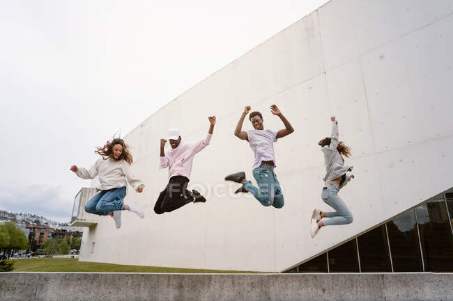 Baixo ângulo de corpo inteiro de amigos multiétnicos levantando as mãos enquanto dá um salto e sorrindo alegremente — Fotografia de Stock