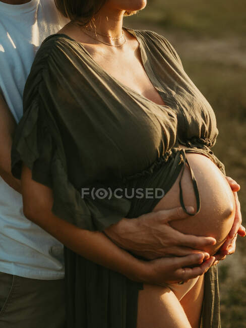 Cropped unrecognizable male hugging pregnant female from behind while standing in countryside meadow — Stock Photo