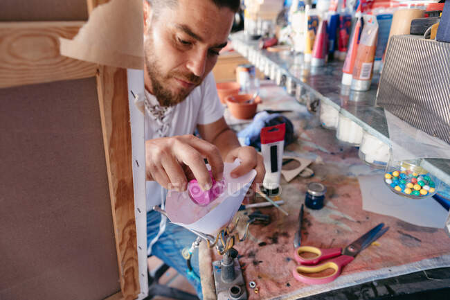 Alto ángulo de artista masculino vertiendo pigmento líquido en pistola de pulverización mientras está sentado cerca de la mesa detrás del caballete con lienzo en el taller - foto de stock