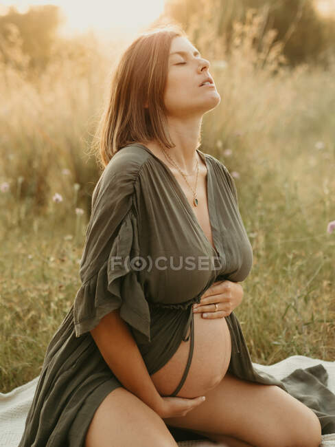 Tranquil pregnant female in dress touching tummy while sitting in field in countryside at sunset in summer — Stock Photo