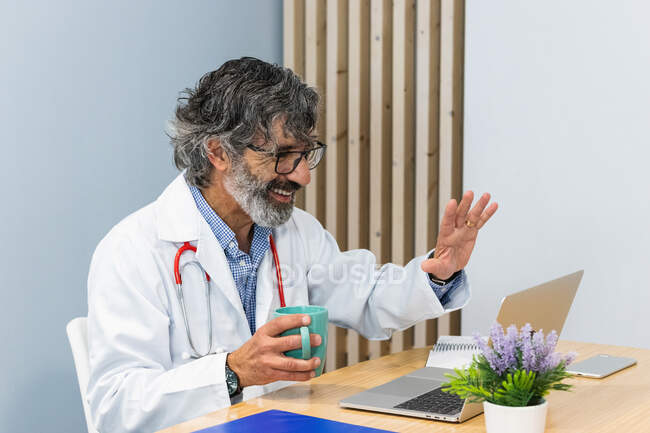 Sonriente médico masculino sénior saludando mano a paciente mientras recibe una videollamada durante la cita por telemedicina a través de una computadora portátil - foto de stock