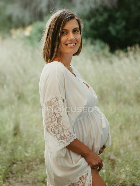 Спокойная беременная женщина в платье трогает животик, стоя в поле в летний день — стоковое фото