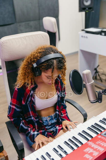 Chanteuse afro-américaine souriante dans un casque jouant du synthétiseur tout en interprétant une chanson en micro en studio d'enregistrement — Photo de stock
