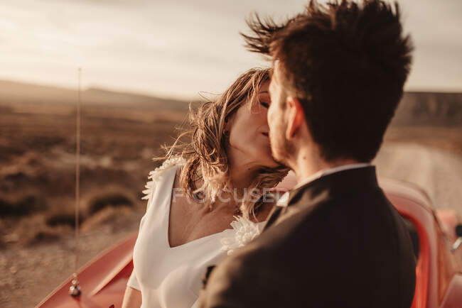 Happy bride and groom embracing and kissing each other while leaning on red luxury vehicle during road trip through Bardenas Reales Natural Park in Navarra, Spain — Stock Photo
