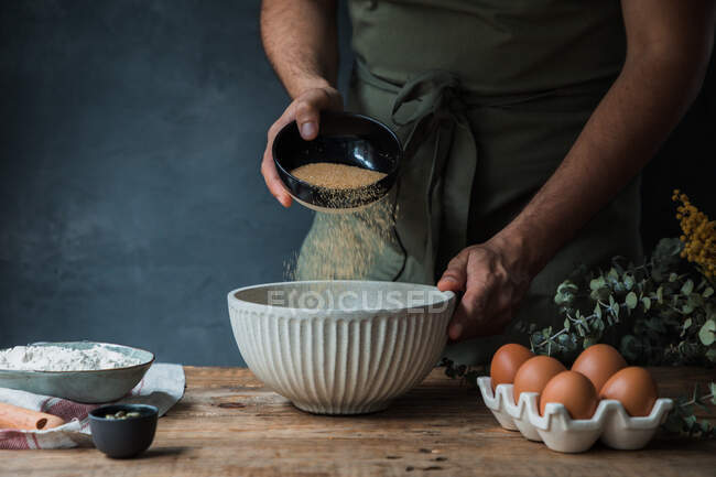Homem irreconhecível em avental derramando açúcar de cana em tigela perto de farinha e ovos enquanto prepara massa para pastelaria — Fotografia de Stock