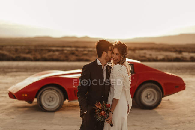 Happy man and woman in elegant clothes standing embracing each other near sports car against sundown sky during wedding celebration in Bardenas Reales Natural Park in Navarra, Spain — Stock Photo