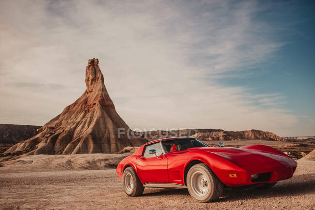 Coche deportivo rojo de lujo aparcado cerca del pico de la montaña contra el cielo nublado en el desierto del Parque Natural de Bardenas Reales en Navarra, España - foto de stock