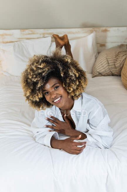 High angle of positive African American female smiling widely while resting on bed and looking at camera — Stock Photo