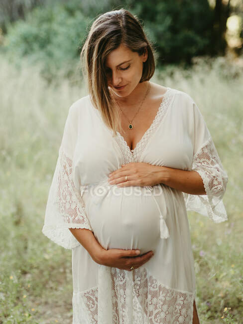 Спокойная беременная женщина в платье касается живота с закрытыми глазами, стоя в поле в сельской местности в летний день — стоковое фото