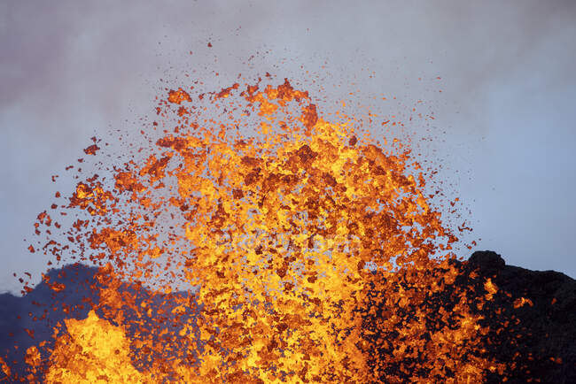 Всплески горячей апельсиновой лавы, извергающейся с вулканической вершины горы, окруженной дымом в Исландии — стоковое фото