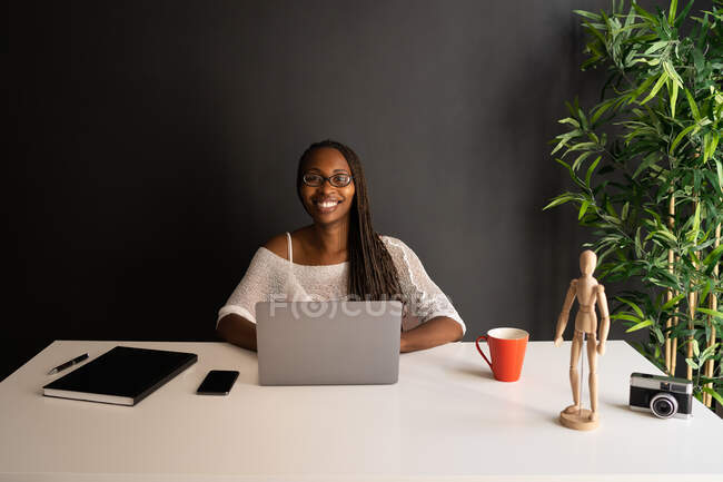 Fröhliche afroamerikanische Freelancerin sitzt am Tisch an einem modernen Arbeitsplatz, während sie fernab von zu Hause an einem Projekt arbeitet — Stockfoto