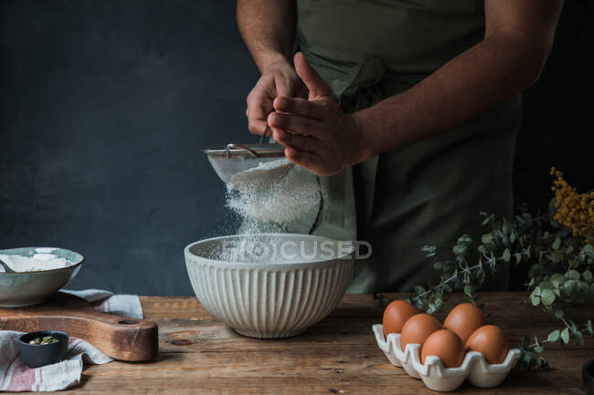 Неузнаваемый человек просеивает муку в миску во время приготовления выпечки на деревенской кухне дома — стоковое фото