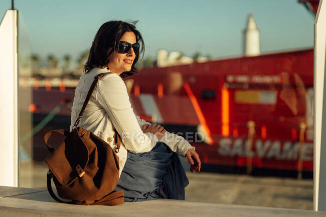 Vista lateral de la mujer viajera adulta sonriente confiada en ropa elegante y gafas de sol con mochila sentada en el banco y mirando hacia otro lado mientras espera el transporte en la ciudad - foto de stock